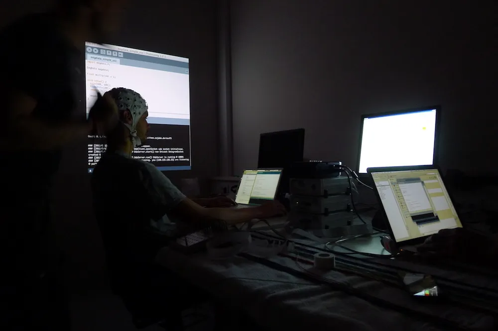 In a dark lab, a man in an EEG electrode cap types at a laptop beneath a projected Processing window titled »eegdata_simple_osc«, surrounded by stacked amplifiers, laptops and a glowing monitor