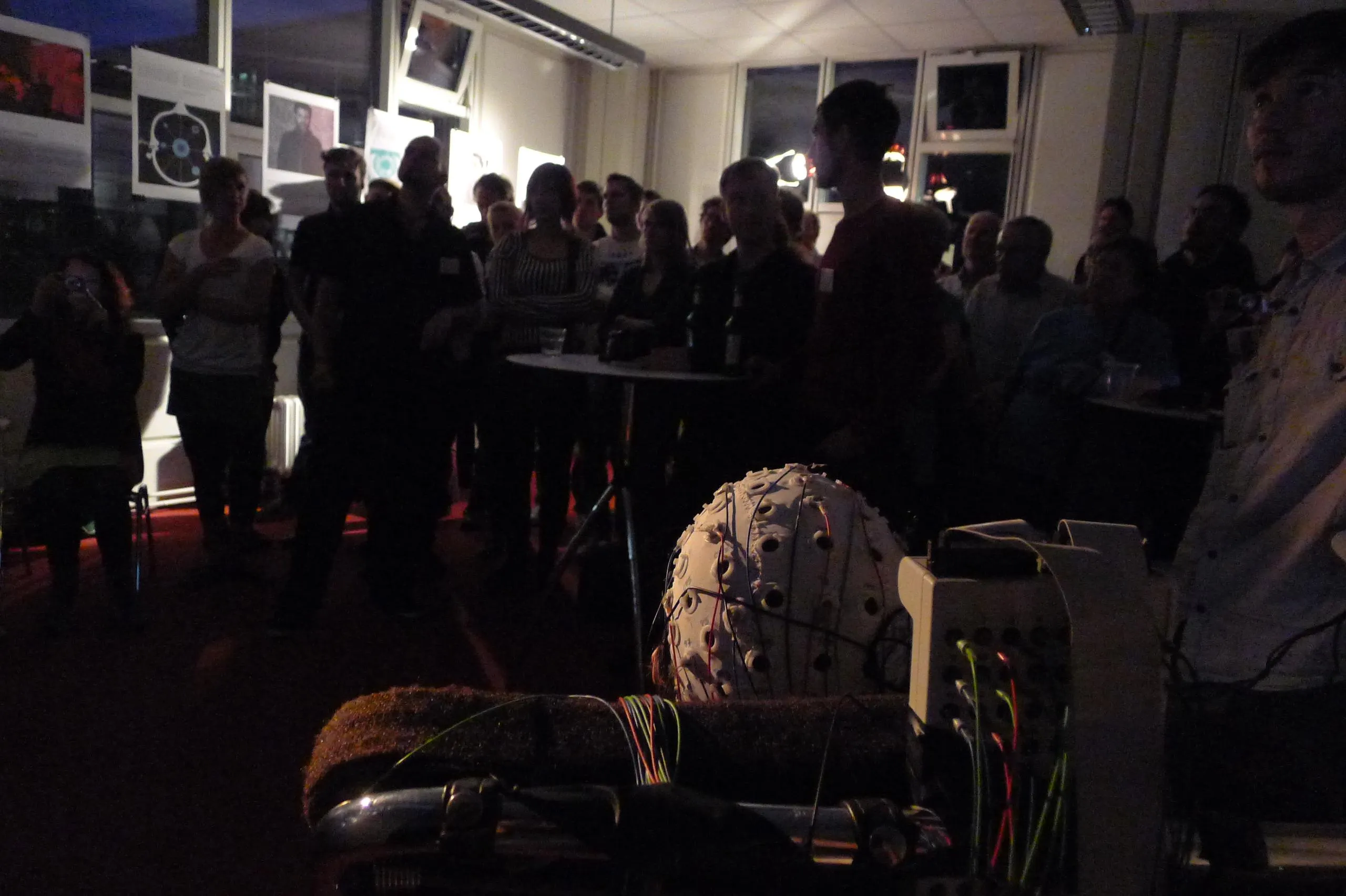 A crowd stands in a dim room hung with posters, watching the experiment; in the foreground, seen from behind, the subject’s head in an EEG electrode cap rests against the chair beside the amplifier with its colored cables