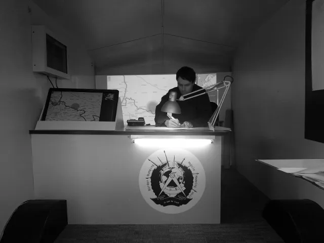 Black-and-white photo of the caravan interior: a man at the desk under the lamp writes on a map, with the touch screen beside him and the projected map behind.