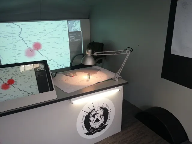 Inside the caravan: a desk with a touch screen and an architect’s lamp over printed maps, in front of a large wall projection of the map with two red incident circles on a border.