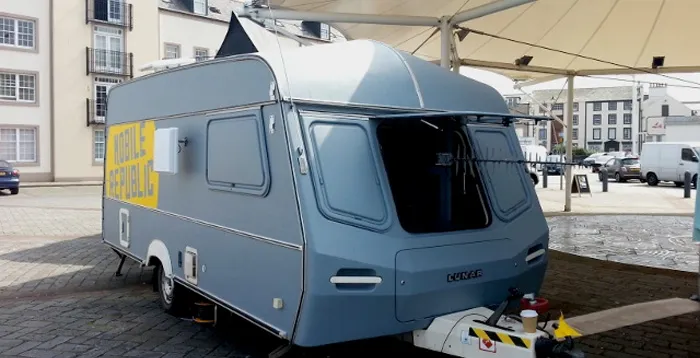 A small gray caravan with a yellow »Mobile Republic« sign on its side, parked under a white tent canopy on a paved square, its front hatch open on a directional antenna.