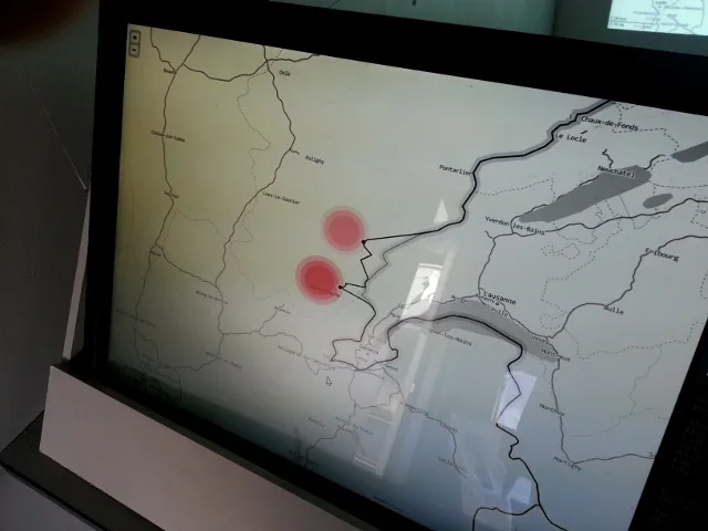The map on the caravan’s touch screen, seen at an angle: two red incident circles on the French–Swiss border in the Jura, northwest of Lake Geneva, where the black border line kinks out toward them.