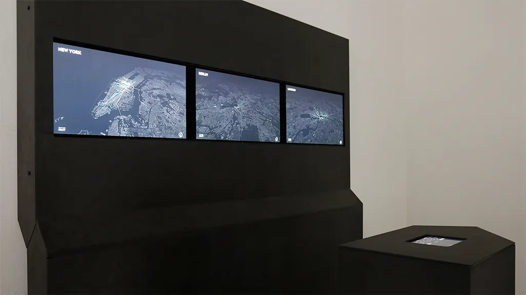 A tall black wall in a white room with three screens set side by side, showing New York, Berlin and London at night from above, and a low black plinth in front with a tablet set into its top