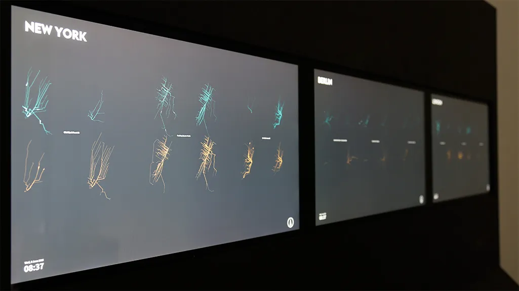 Close view of the three screens: New York in front shows six stations as pairs of trip-trail glyphs, cyan above and orange below with the station name between them, while Berlin and London recede into the dark