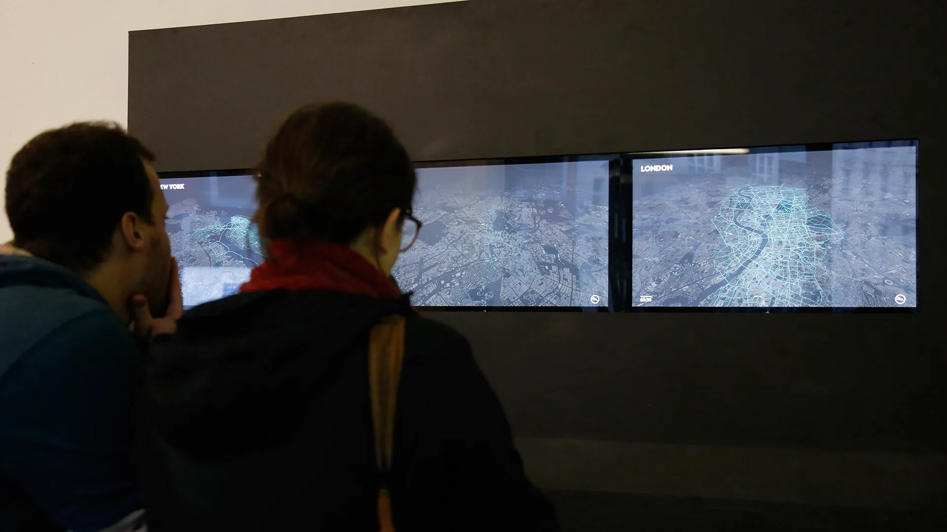 Two visitors seen from behind, a man with a hand to his chin and a woman with glasses and a red scarf, studying the three screens, where London’s trails glow cyan over the gray city map