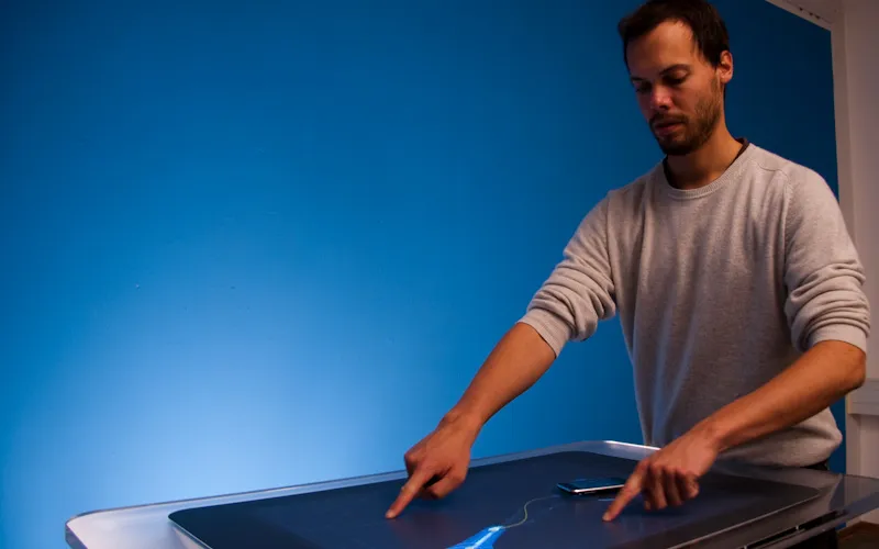 Christopher Pietsch stands at the Microsoft Surface table against a saturated blue wall, touching the tabletop with both index fingers; a smartphone lies on the surface beside a thin blue trace of liquid