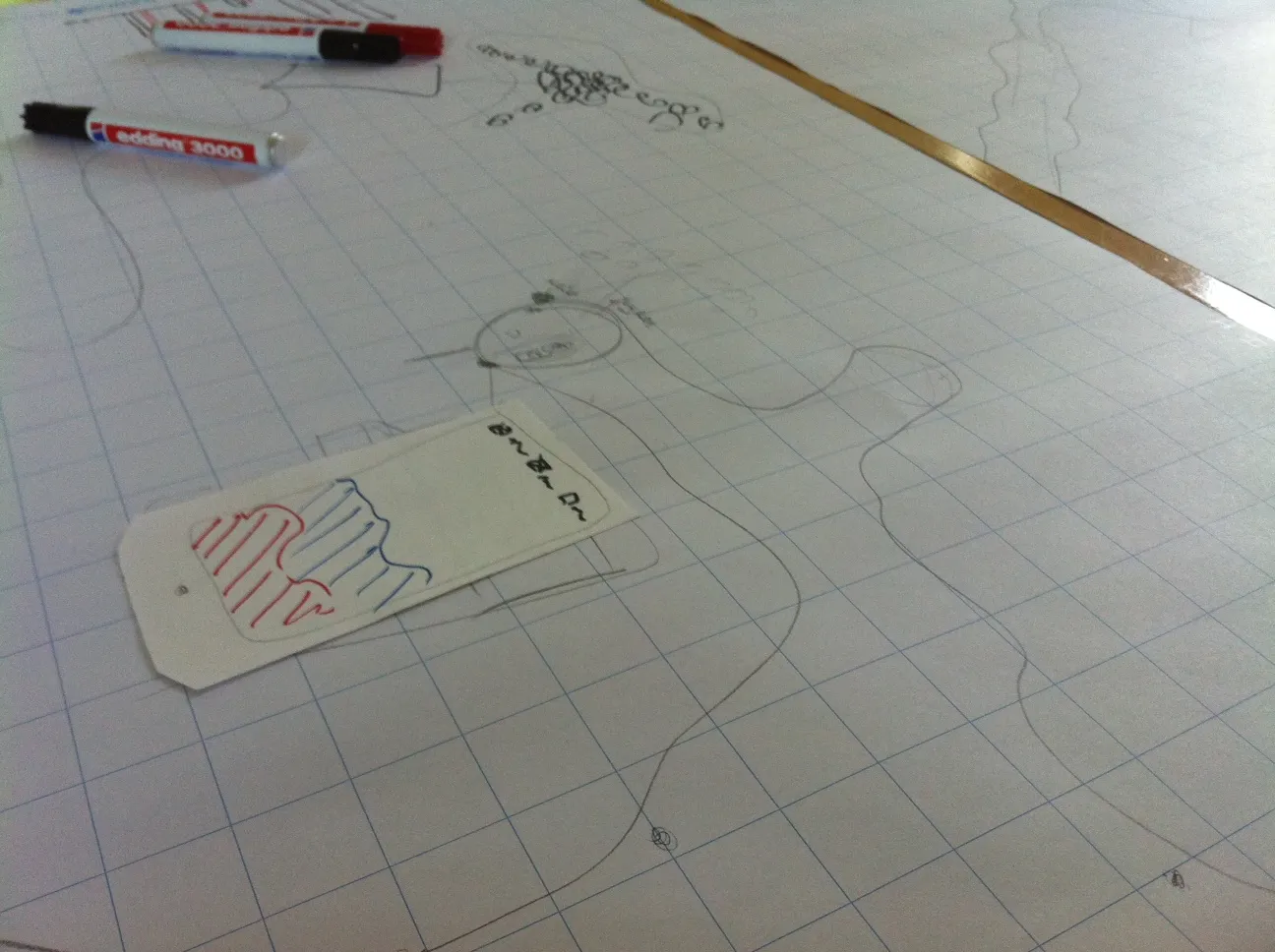 A large sheet of grid paper with pencil-drawn streets and loops and a paper phone cut-out hatched in red and blue, with several felt-tip markers lying at the top edge