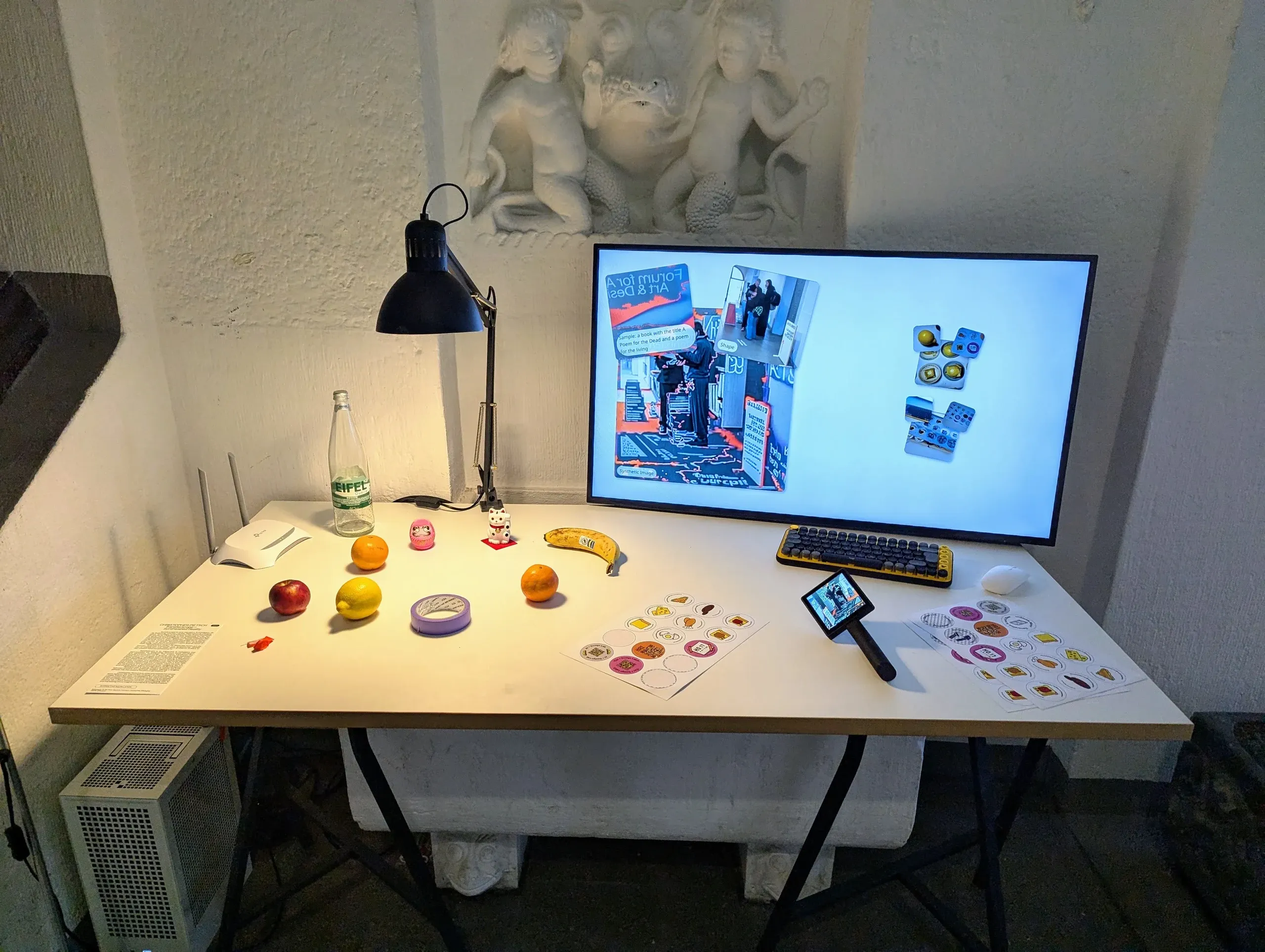 A white table set up for sampling: a desk lamp lights two oranges, a lemon, an apple, a banana and a water bottle among sticker sheets, while Transferscope lies in front of a monitor showing a sample, its shape and the synthetic image.