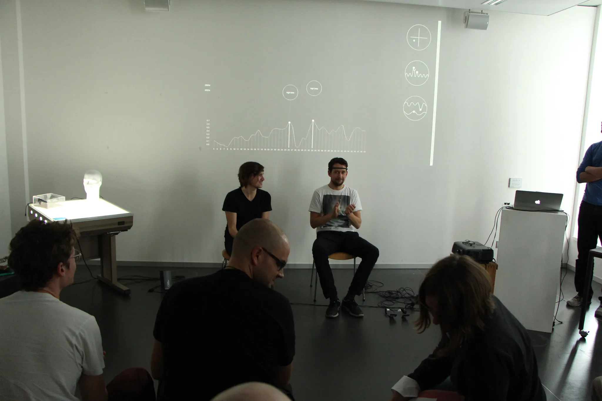 An audience sits facing two men on chairs beneath the projected frequency curve with its two “high beta” peaks; a laptop on a white plinth stands at right, and a white head on a glowing light table beside a clear acrylic box at left