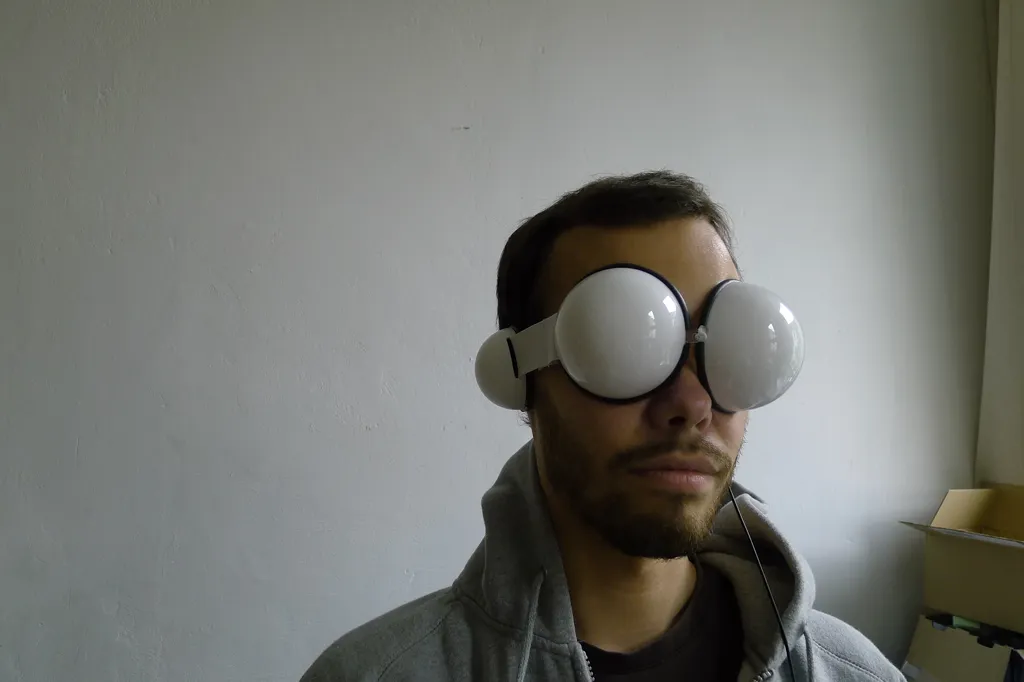 A man in a grey hoodie wears a diffuser mask: two white plastic hemispheres over his eyes and a smaller one over his ear as a headphone shell, joined by a white strap