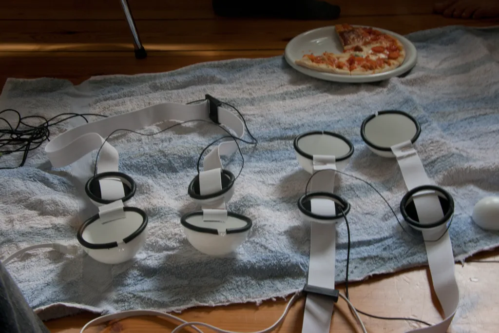Four finished diffuser masks lie on a towel, each a pair of white hemispheres edged with black rubber and joined by white straps and cables, next to a plate of pizza
