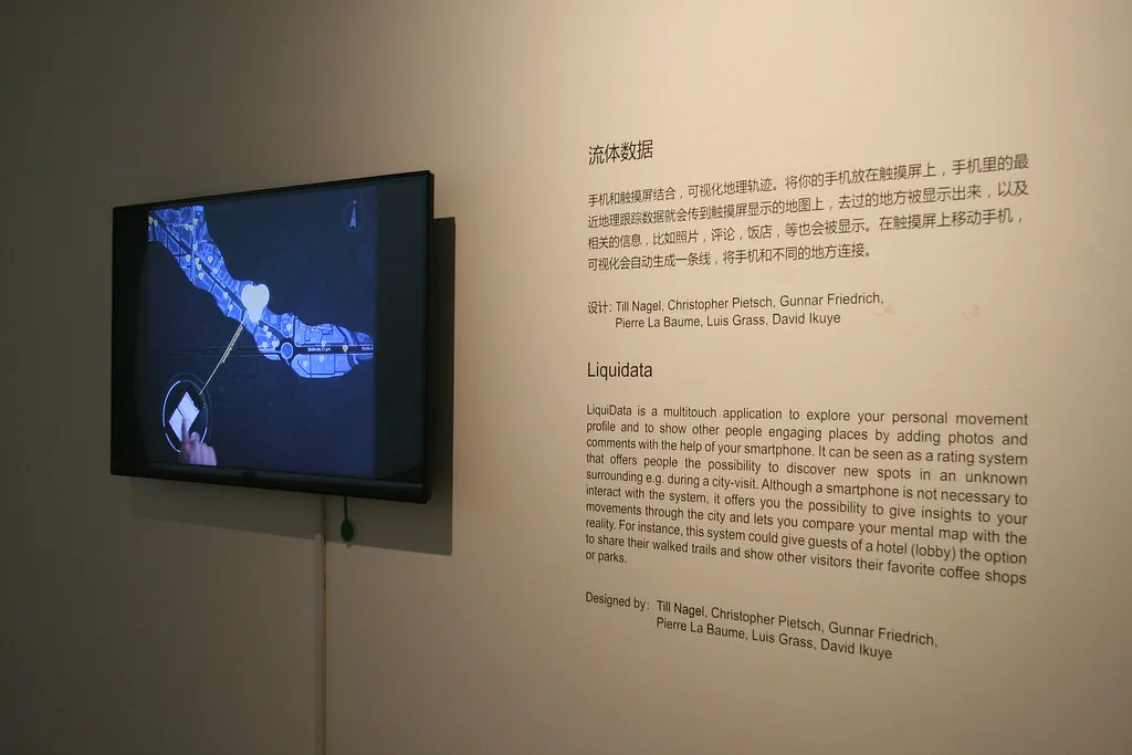 A wall-mounted screen plays the LiquiData video, a phone in its ring tied to a place on the blue map, beside wall text in Chinese and English headed »流体数据« and »Liquidata« that credits Till Nagel, Christopher Pietsch, Gunnar Friedrich, Pierre La Baume, Luis Grass and David Ikuye