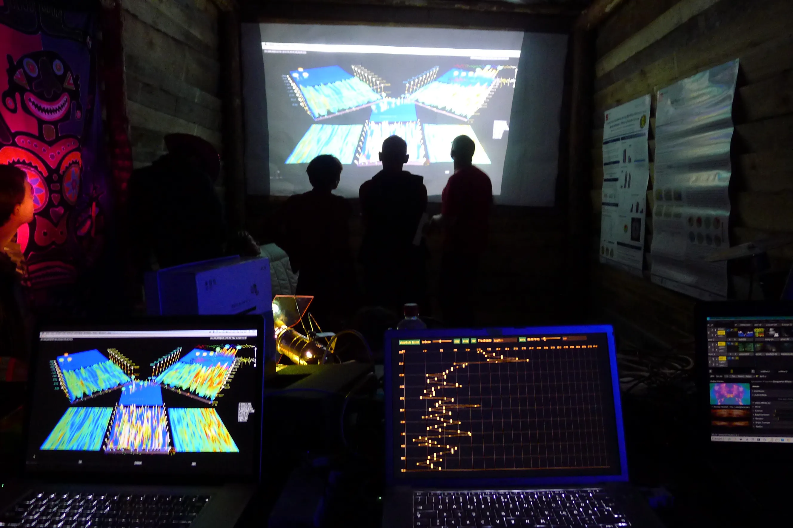 In a dark wooden hut hung with posters and a bright patterned cloth, silhouetted visitors stand before a projected 3D landscape of EEG data; in the foreground one laptop mirrors the landscape and another shows an orange session curve