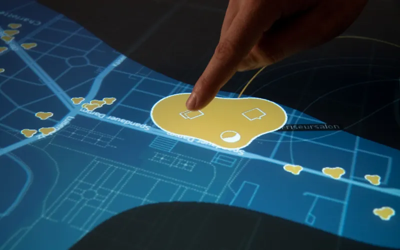 A fingertip touches a place opened into a large yellow blob with three small white icons on a blue street, smaller yellow place markers scattered along the streets around it
