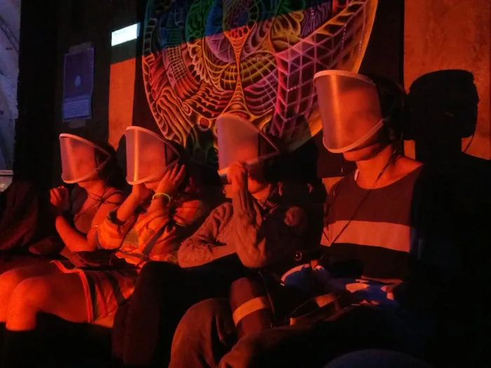 Four people recline side by side on a bench in red light, each wearing a translucent visor over the face, in front of a large psychedelic wall hanging