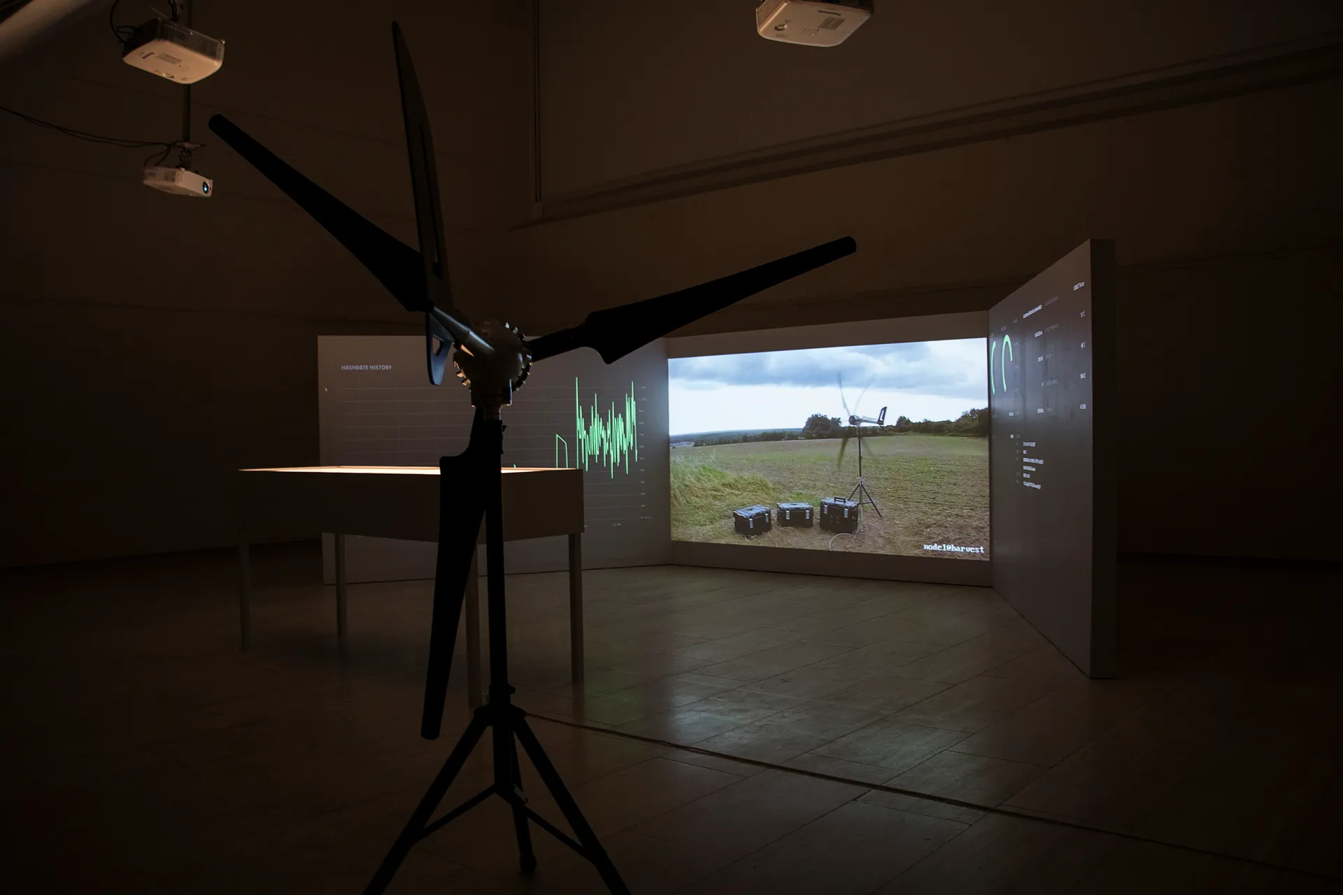The turbine on its tripod in the darkened gallery, in front of three projection walls: the hashrate history on the left, a live view of the turbine in its field in the middle, and the dashboard figures on the right, with the lit vitrine to one side.