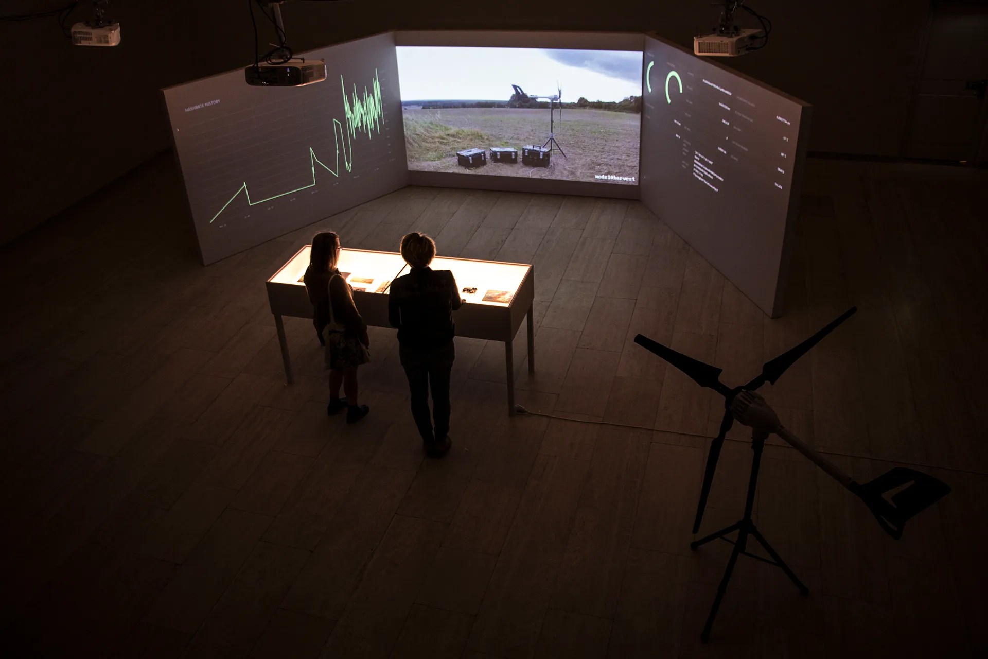 Seen from above, two visitors stand at the lit vitrine facing the three projection walls, with the turbine on its tripod in the foreground.