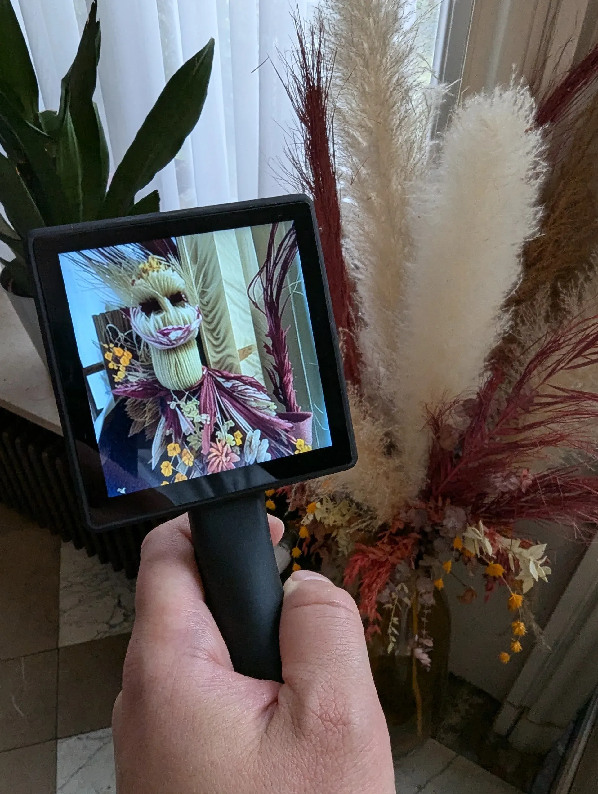 A hand holds Transferscope beside a tall arrangement of pampas grass and dried flowers; its screen shows a figure whose head and body are made of the same grasses and blossoms.