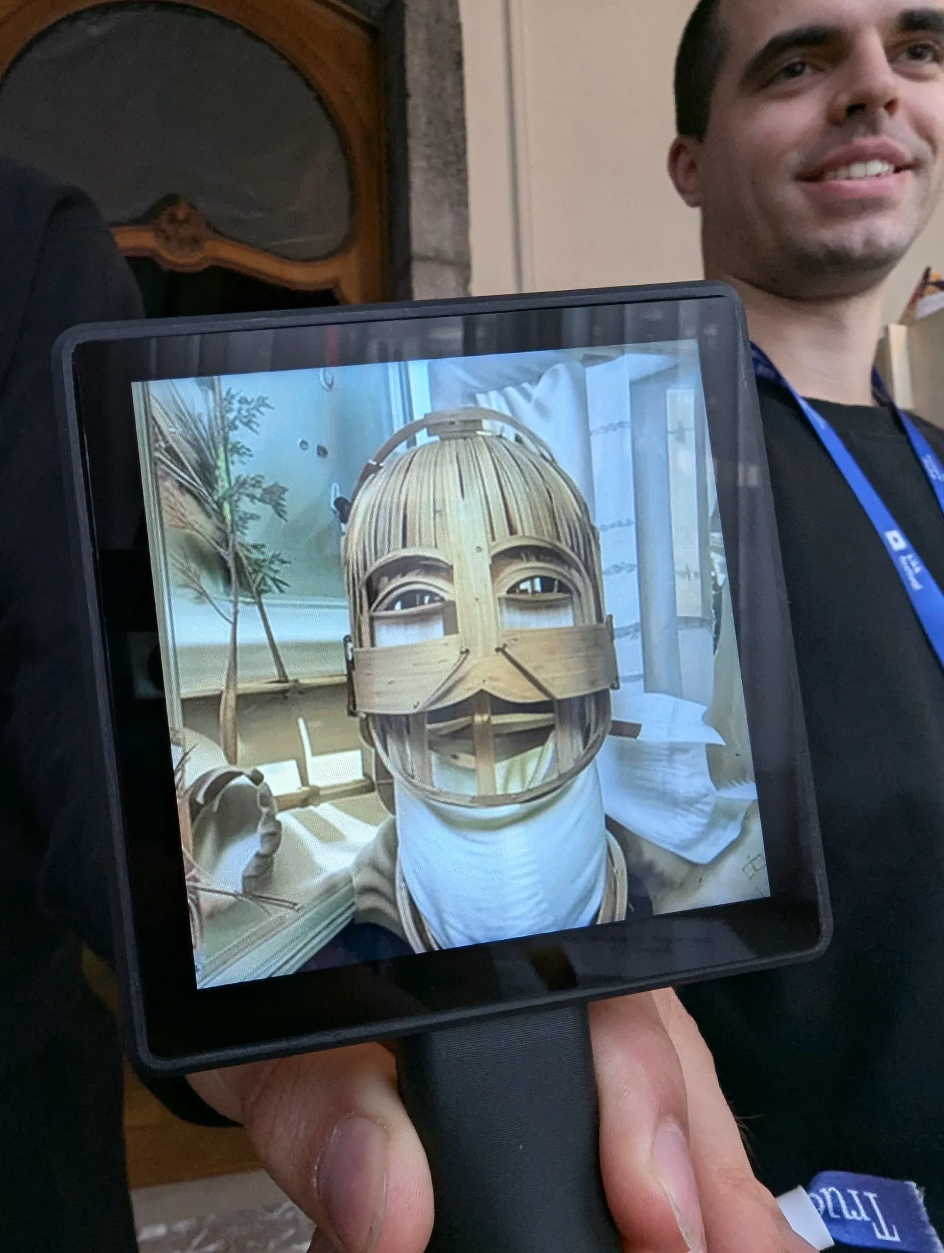 Transferscope held up in front of a smiling visitor with a festival lanyard; its screen shows a face rebuilt as a woven cage of thin wooden strips above a white collar.