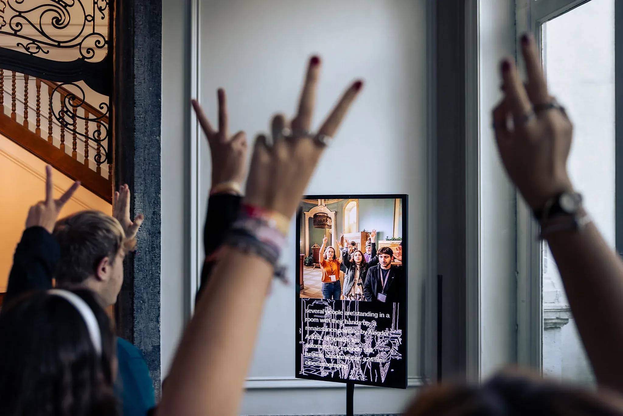 Visitors raise peace signs toward unStable Mirror; on its upper screen they reappear with raised hands in an ornate room, and below, the edge map carries the caption “several people are standing in a room with their hands up, a renaissance painting by Avigdor Arikha …”