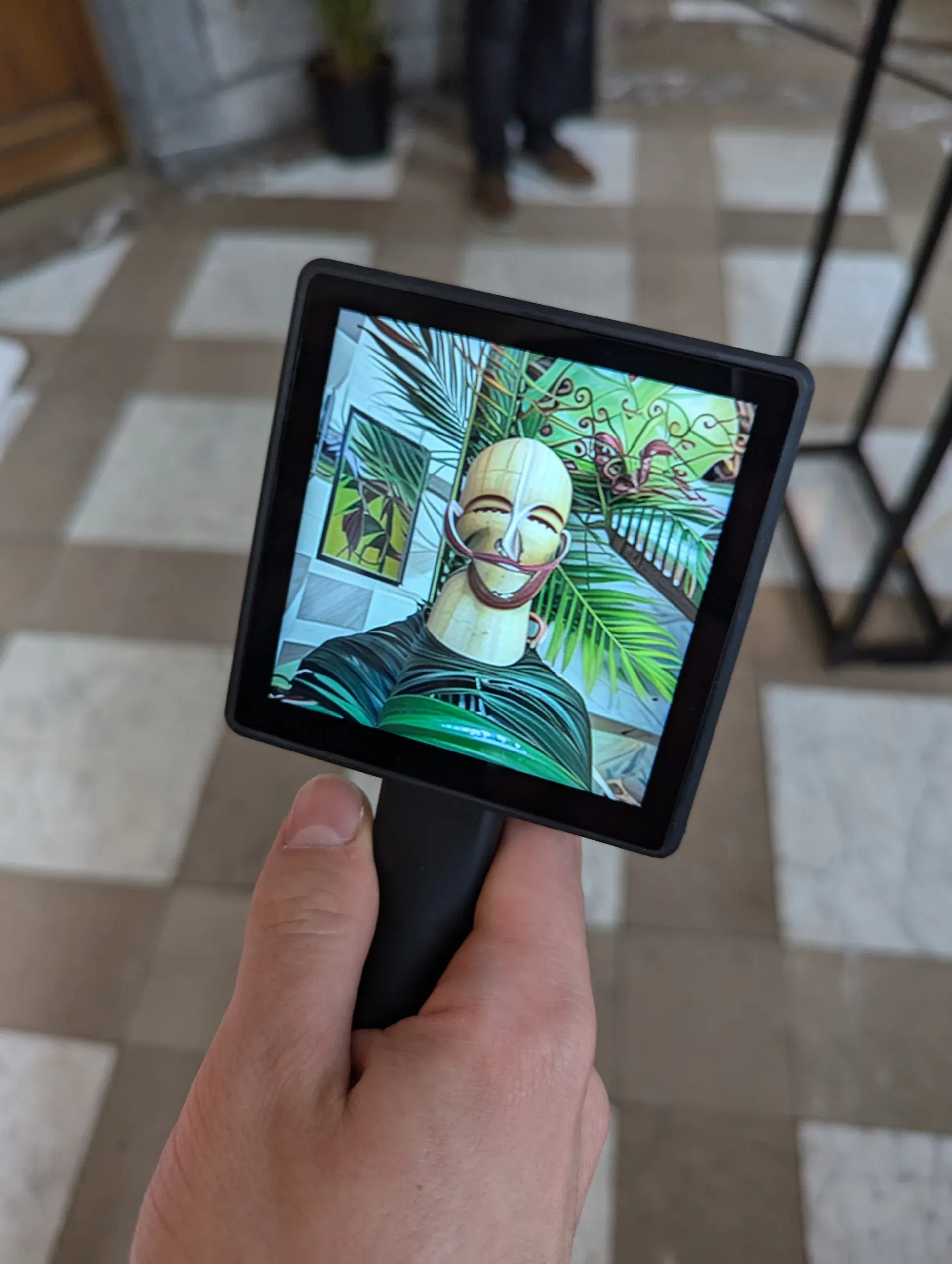 A hand holds Transferscope above a checkered stone floor; its screen shows a person reimagined as a pale wooden figure with a carved, mask-like face, framed by palm leaves.