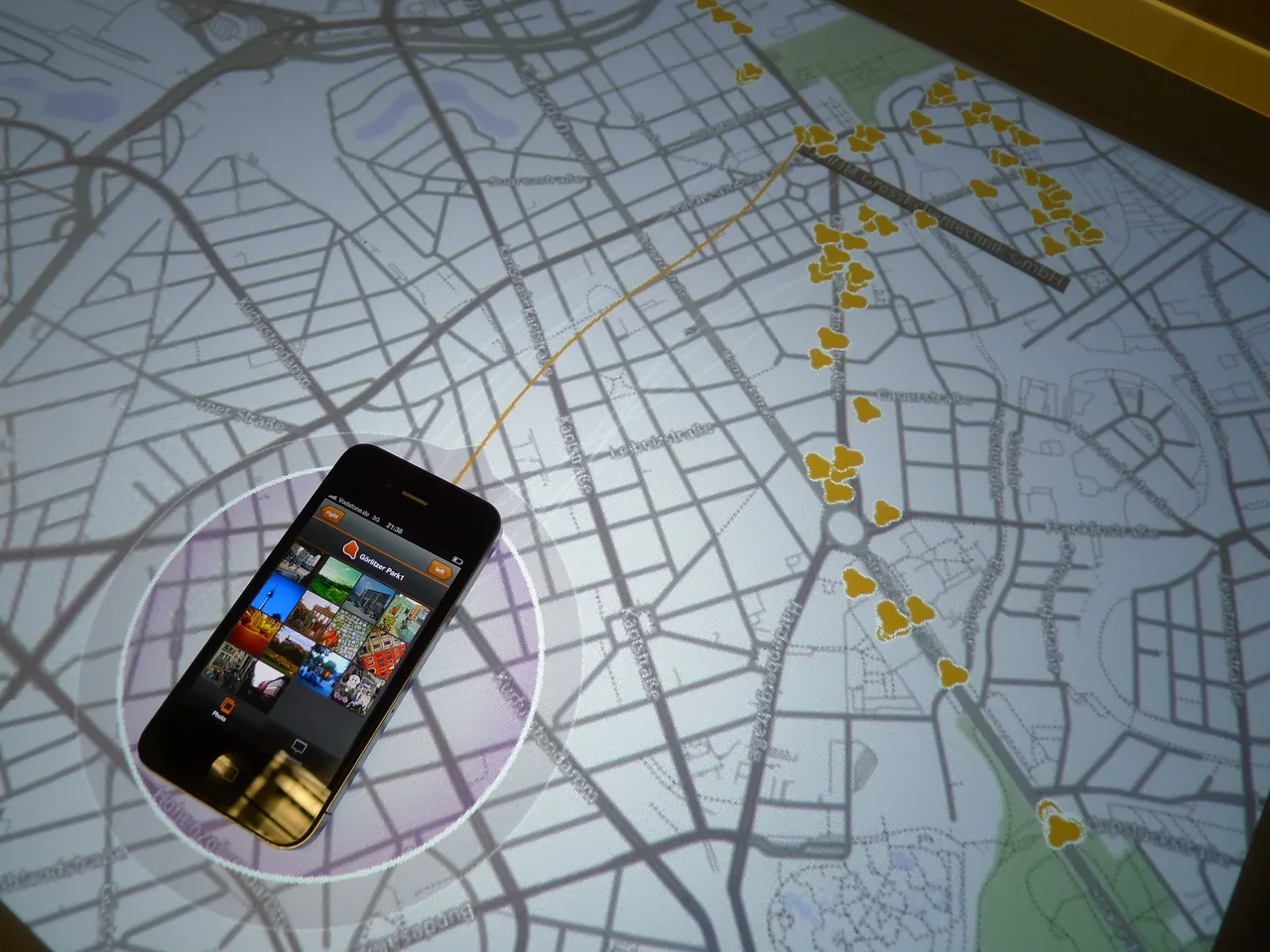 A phone showing a grid of photos for »Görlitzer Park1« lies in a purple-tinted ring on a light gray street map, an orange line running from it to long clusters of yellow place markers