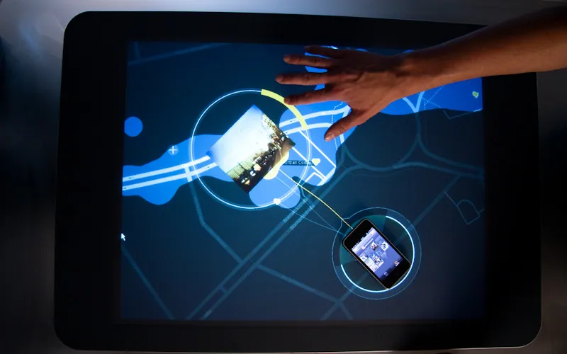 The whole tabletop seen from above: a band of blue liquid crosses the dark street map, a photo opens inside a circle with a yellow arc, and a phone in its glowing ring is tied to it by a thin yellow line while a hand reaches across the surface