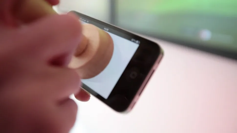 A hand presses the round wooden stamp onto the screen of an iPhone, seen at an angle against a soft green and pink background