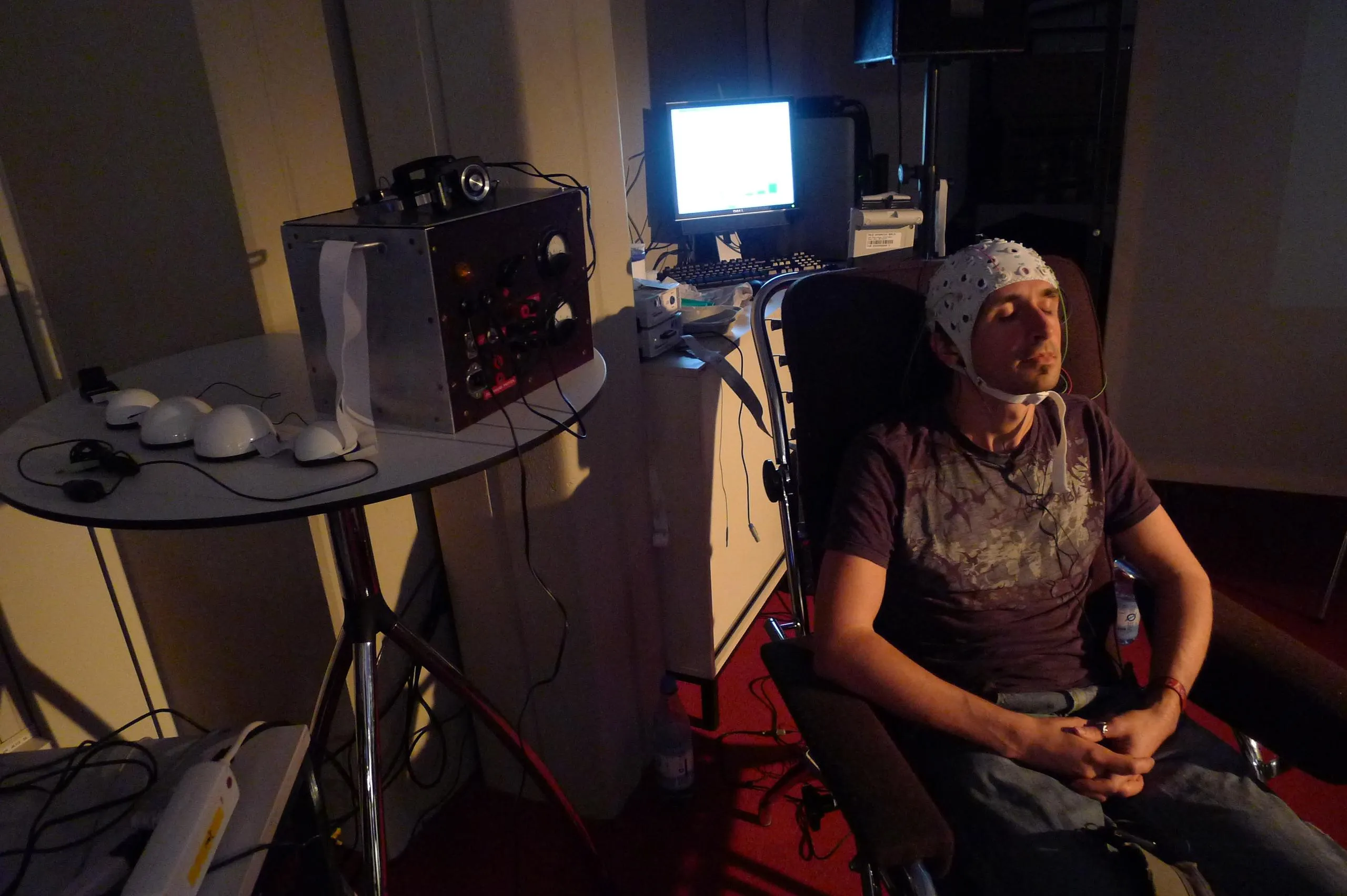 A man in a white EEG electrode cap reclines with eyes closed in a dark chair; beside him, a round table holds four white plastic hemispheres and the metal box with dials and gauges that served as the fake control panel, a pair of headphones on top