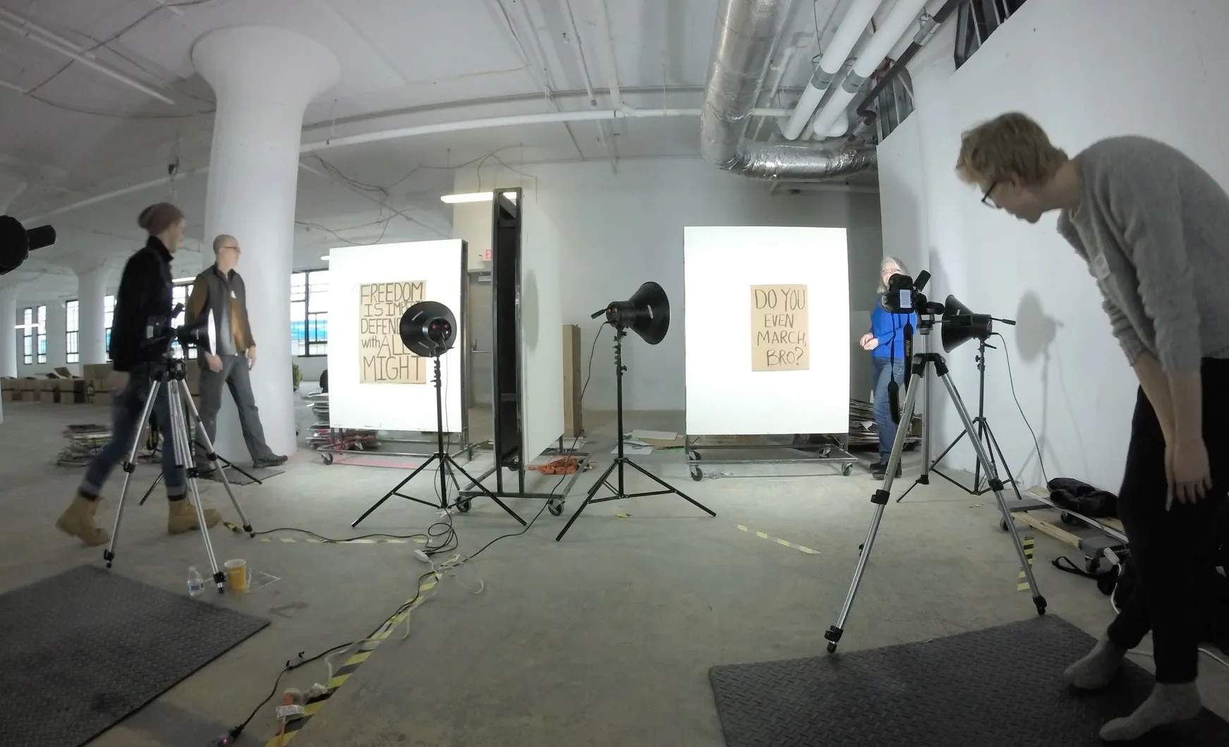 Two makeshift photo stations in the loft: cardboard signs, one partly hidden behind a flash and ending »…with all might«, the other »Do You Even March, Bro?«, pinned to white boards on wheels and lit by studio flashes, with cameras on tripods and volunteers checking the shots