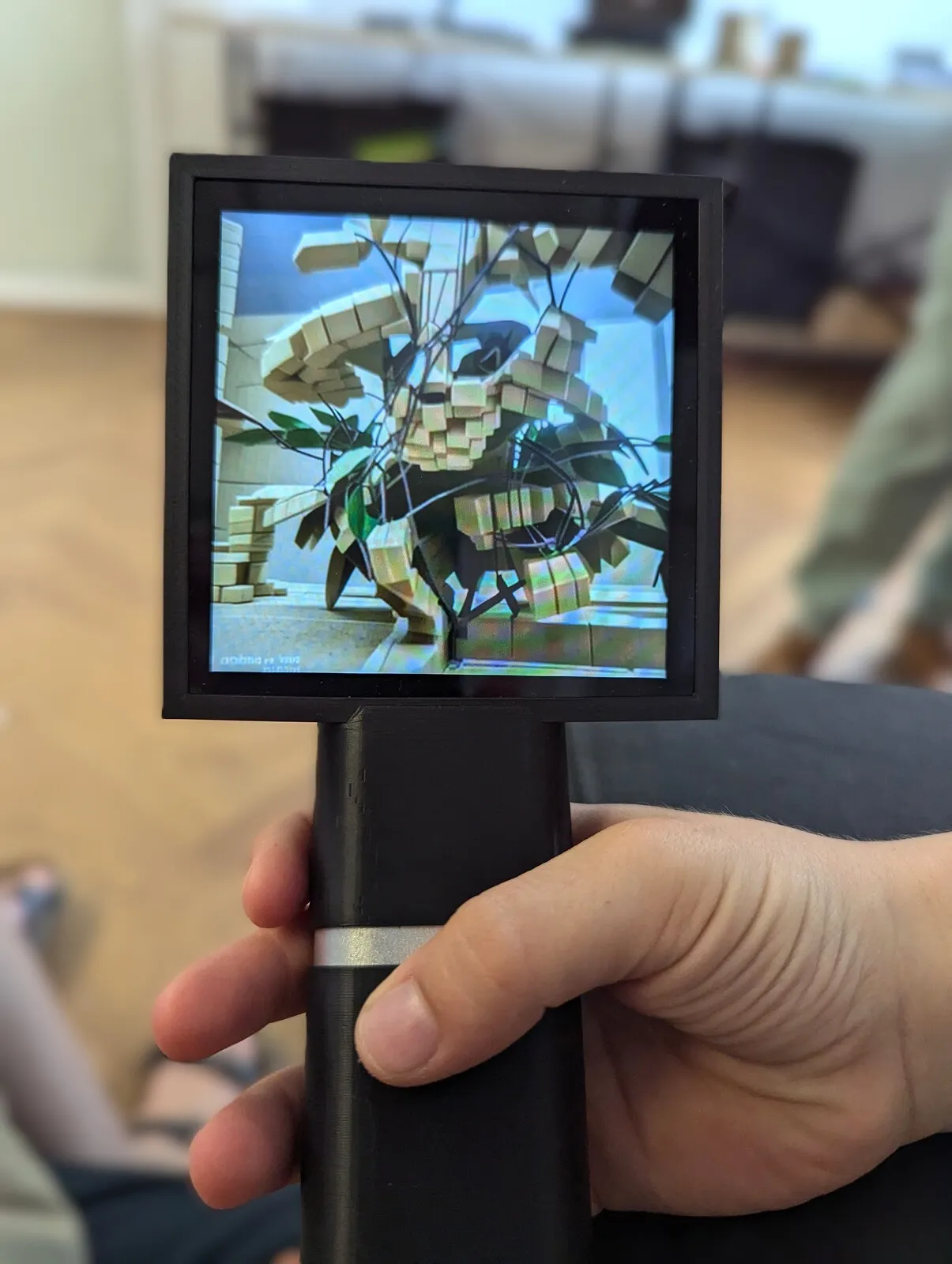Transferscope held in a hand, its screen showing a potted plant rebuilt from stacked light wooden blocks among dark leaves.