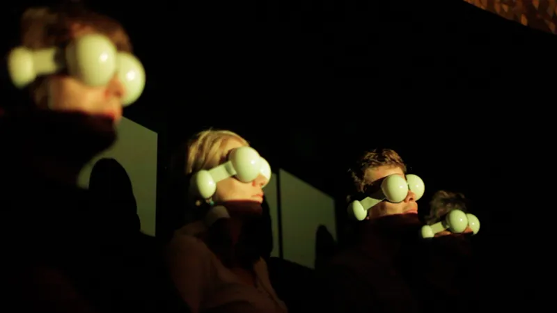 Four volunteers sit in a row in the dark, each wearing a diffuser mask with two pale spheres over the eyes and a smaller one at the ear, their faces lit warmly from the side