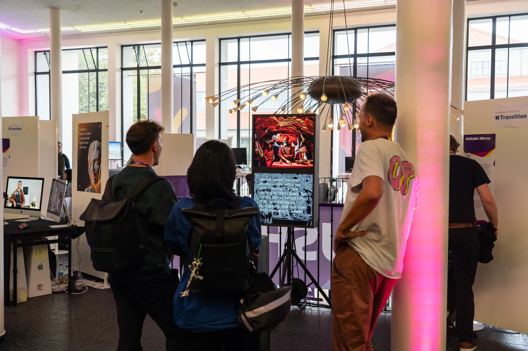 Conference visitors with backpacks stand in front of the mirror in a bright hall with tall windows and a spiky chandelier; the screens show a generated painting of people at a table above its edge map, an “unStable Mirror” sign on the wall behind
