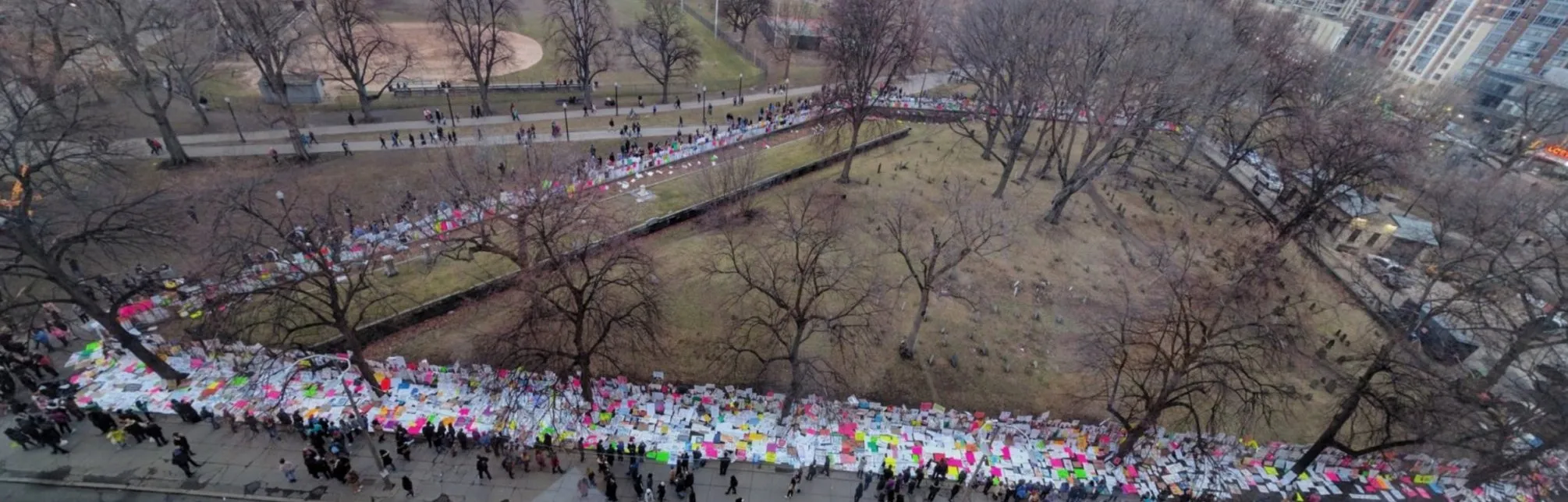 A wide panorama from high above Boston Common: thousands of colorful protest signs lie in a long band along the fence of the Central Burying Ground, between bare trees and old gravestones, while marchers stream along the sidewalk and the paths beyond