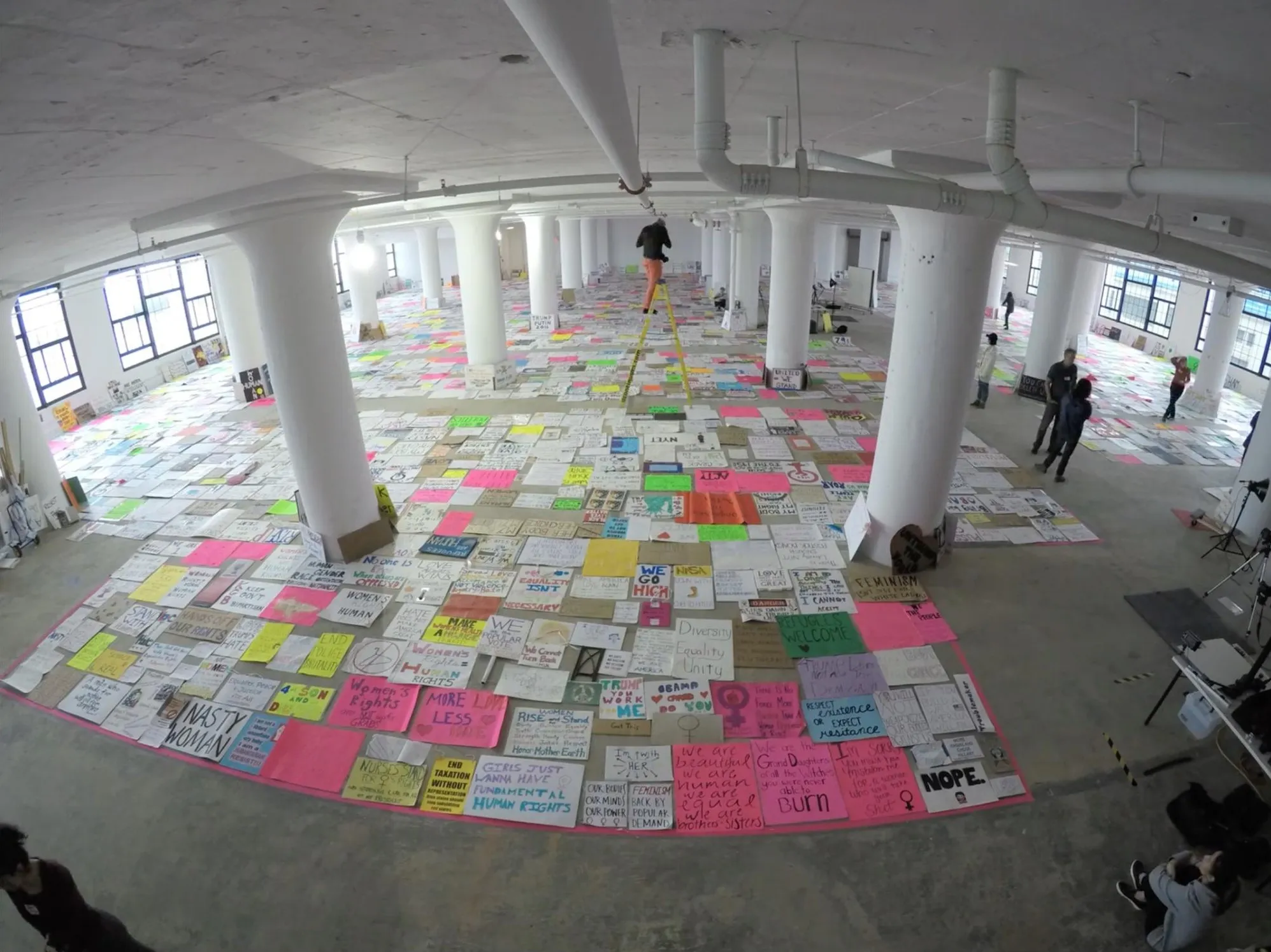 A fisheye view across a large white loft with columns, its whole floor covered in rows of protest signs laid out side by side, »Nasty Woman«, »Refugees Welcome« and »Nope.« among them, while a volunteer stands on a yellow ladder in the middle