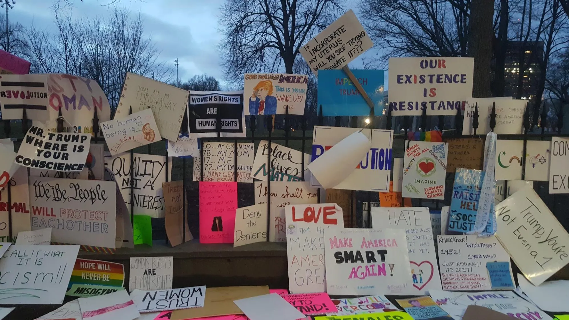 Handmade signs crowd the iron fence at dusk, stacked and overlapping: »Our Existence Is Resistance«, »Mr. Trump Where Is Your Conscience«, »We the People Will Protect Each Other«, »Make America Smart Again!« and a hand-drawn Trump cartoon under »Good Morning America«