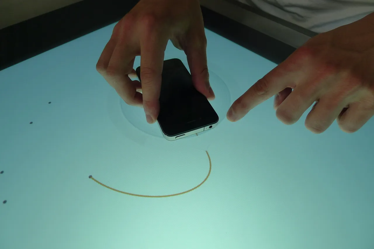Two hands on a bright, nearly empty tabletop: one holds a phone inside a faint circle, the other points beside it, and a thin orange curved line arcs away from the phone