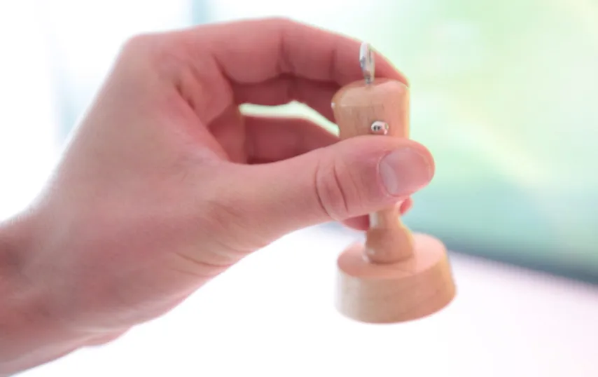 A hand holds the small turned-wood stamp by its handle, a metal eyelet on top and a small metal stud on its side, against a bright blurred background