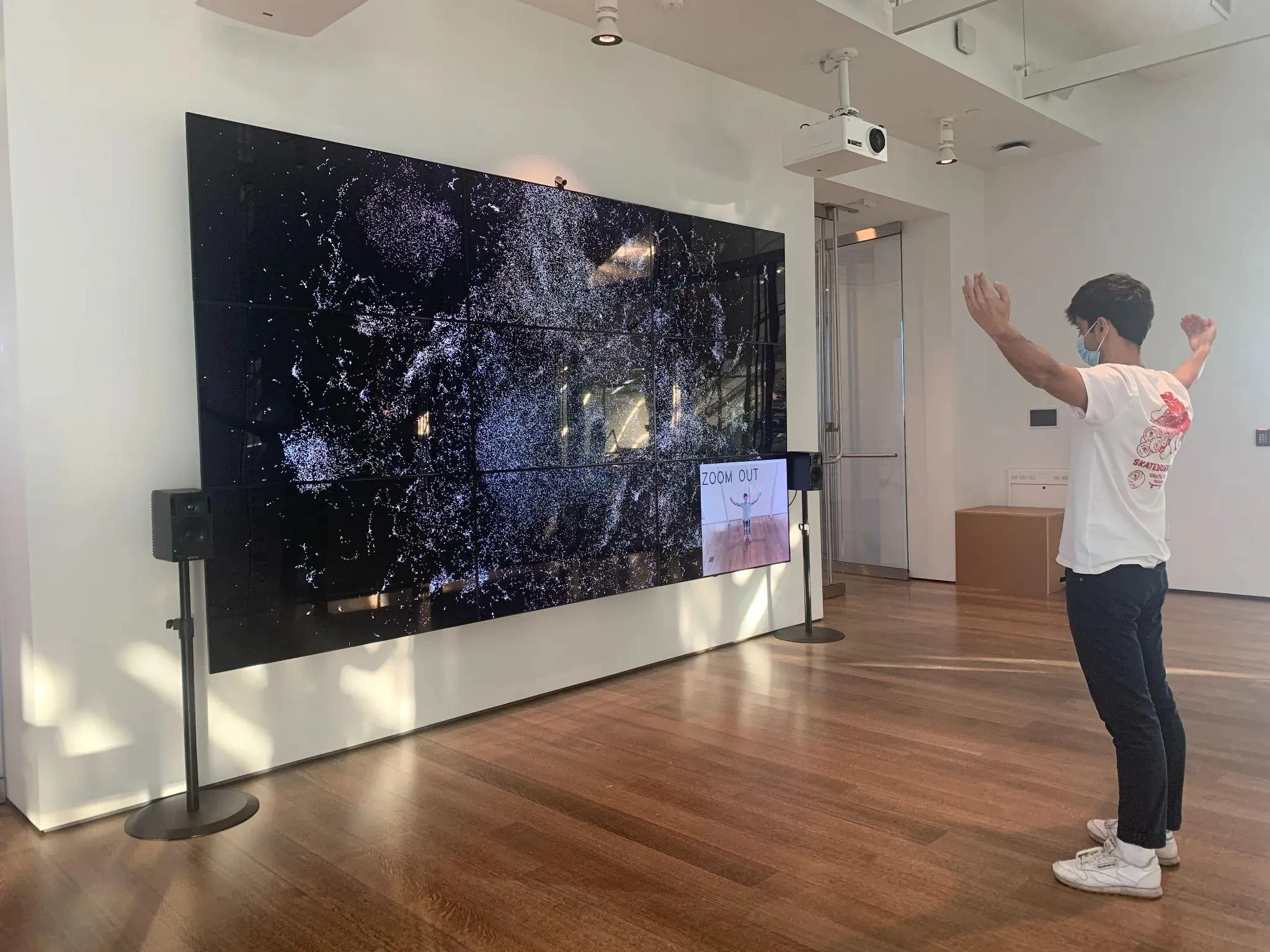 A man in a white T-shirt with a red print on the back and a face mask stands on a wooden floor with both arms raised before the nine-monitor wall, which shows the collection as a field of white points; a small screen at the wall’s lower right shows the camera image of his pose under the words ZOOM OUT