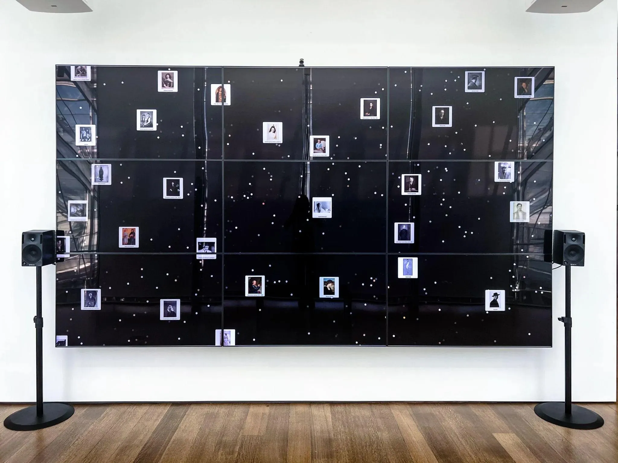 The Lightbox Gallery’s wall of nine monitors in Trails mode, zoomed in: a black field of white dots standing in for the collection’s objects, with a few dozen sampled portraits shown as small white-framed cards with captions, flanked by two speakers on stands