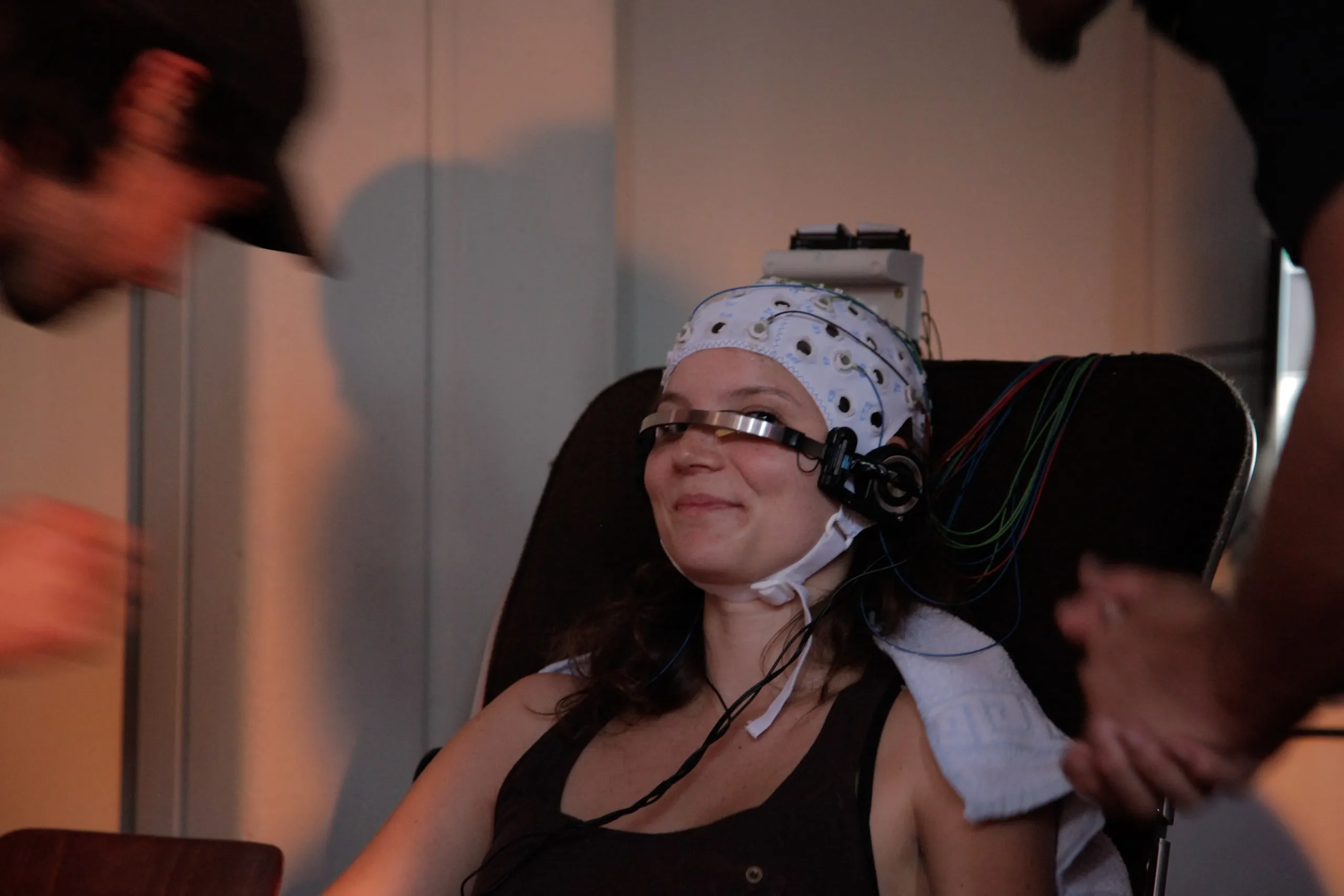 A smiling woman in a white EEG electrode cap leans back in a chair, the brain machine’s metal band across her eyes and a headphone at her ear, while two blurred helpers lean in from the edges of the frame
