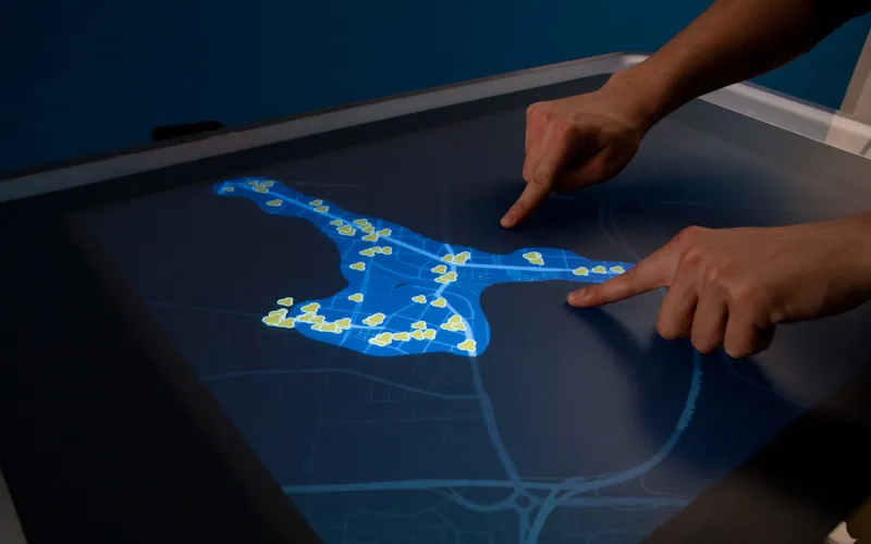Two index fingers trace the bright blue liquid on the dark tabletop map, its streets dotted with small yellow place markers
