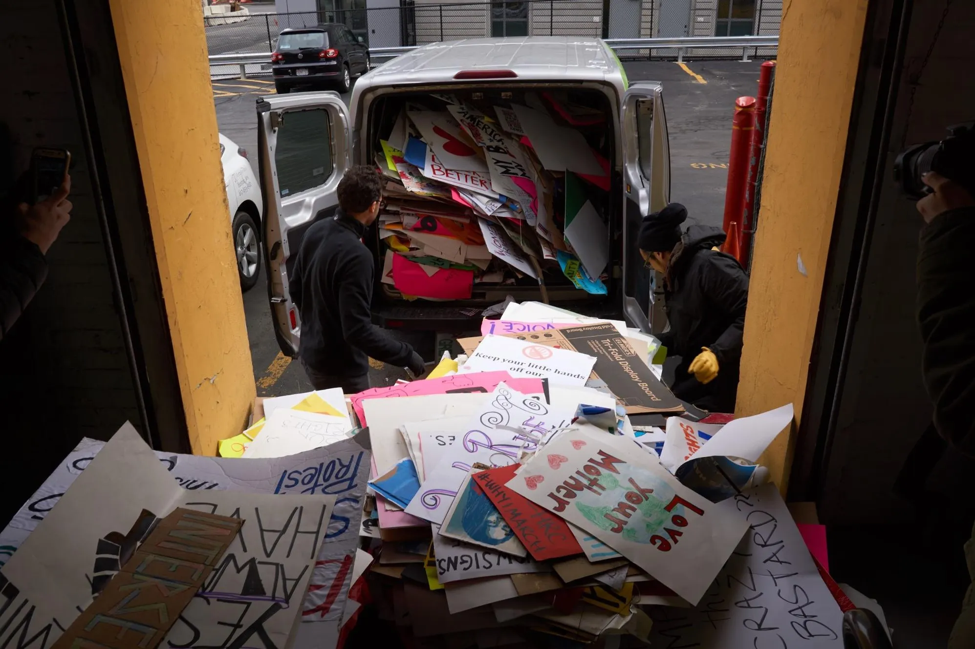 Seen from inside a loading dock between two yellow door posts, two people unload the overflowing van onto a heap of signs in the foreground, among them »Love Your Mother« with a drawn globe and »#WhyIMarch«