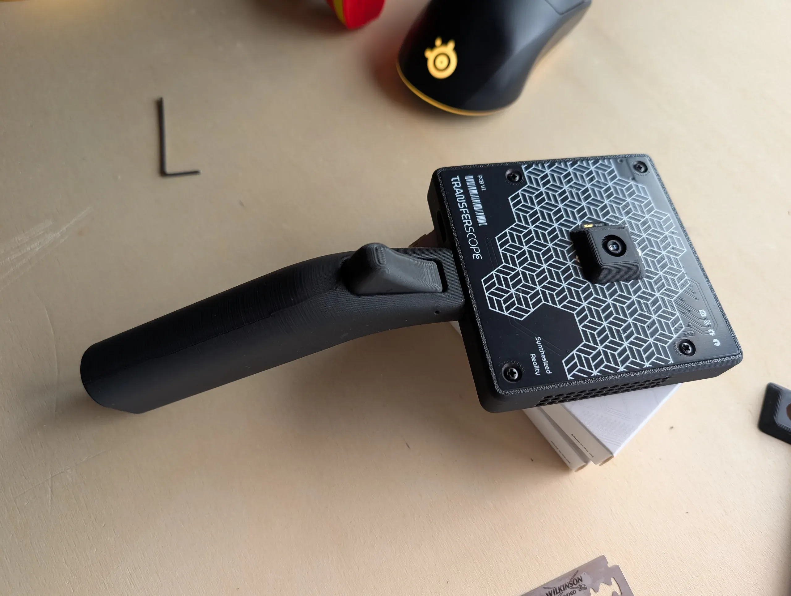 The back of Transferscope version 2: a black circuit board printed with a white cube pattern and the words “Transferscope” and “Synthesized Reality” forms the back panel, with the camera module in its center and the curved grip with its trigger beside it