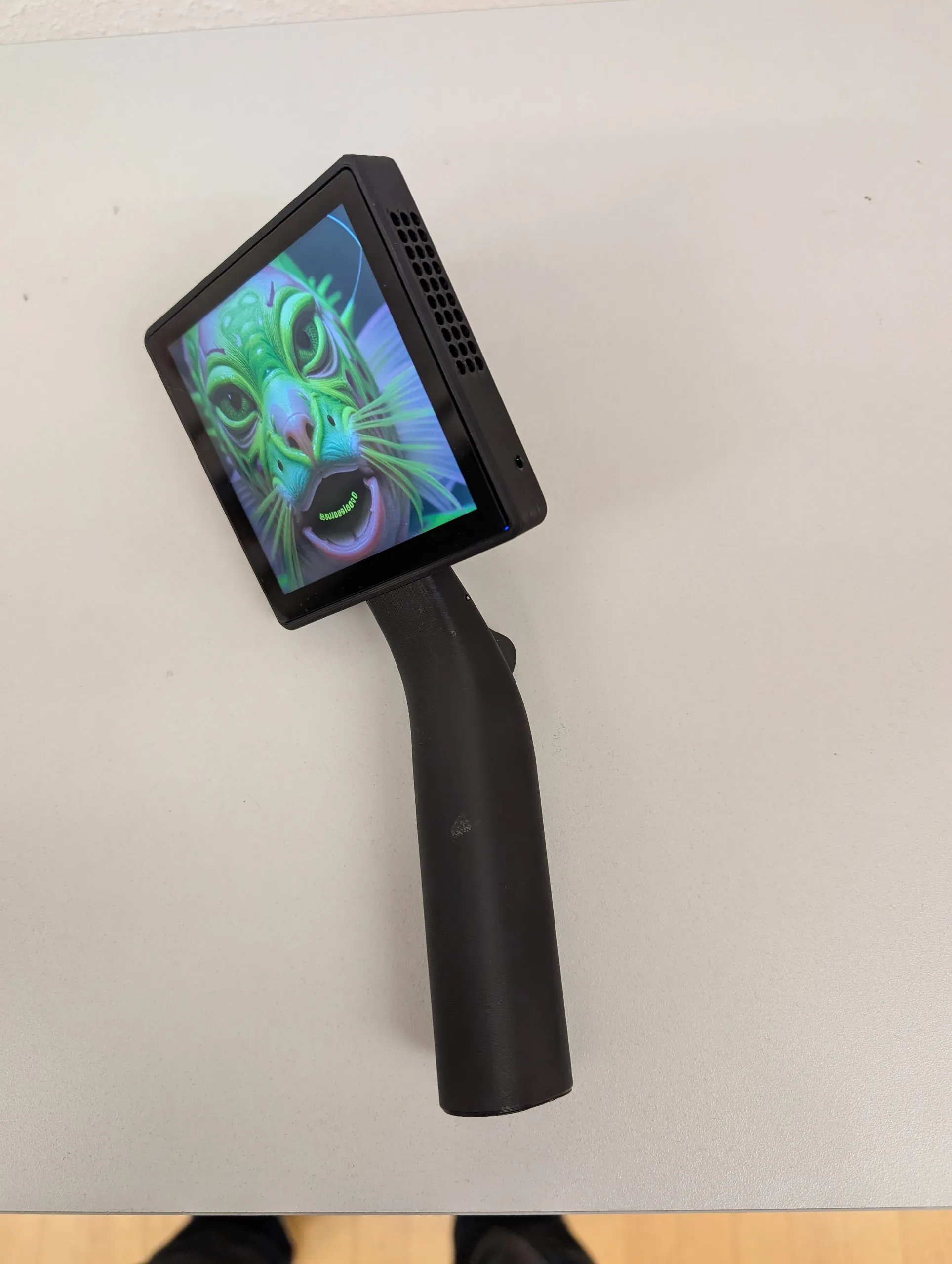 The second version of Transferscope lying on a white table, with its curved grip and trigger; the square screen shows a synthesized creature with a green, fish-like face and a wide-open mouth.