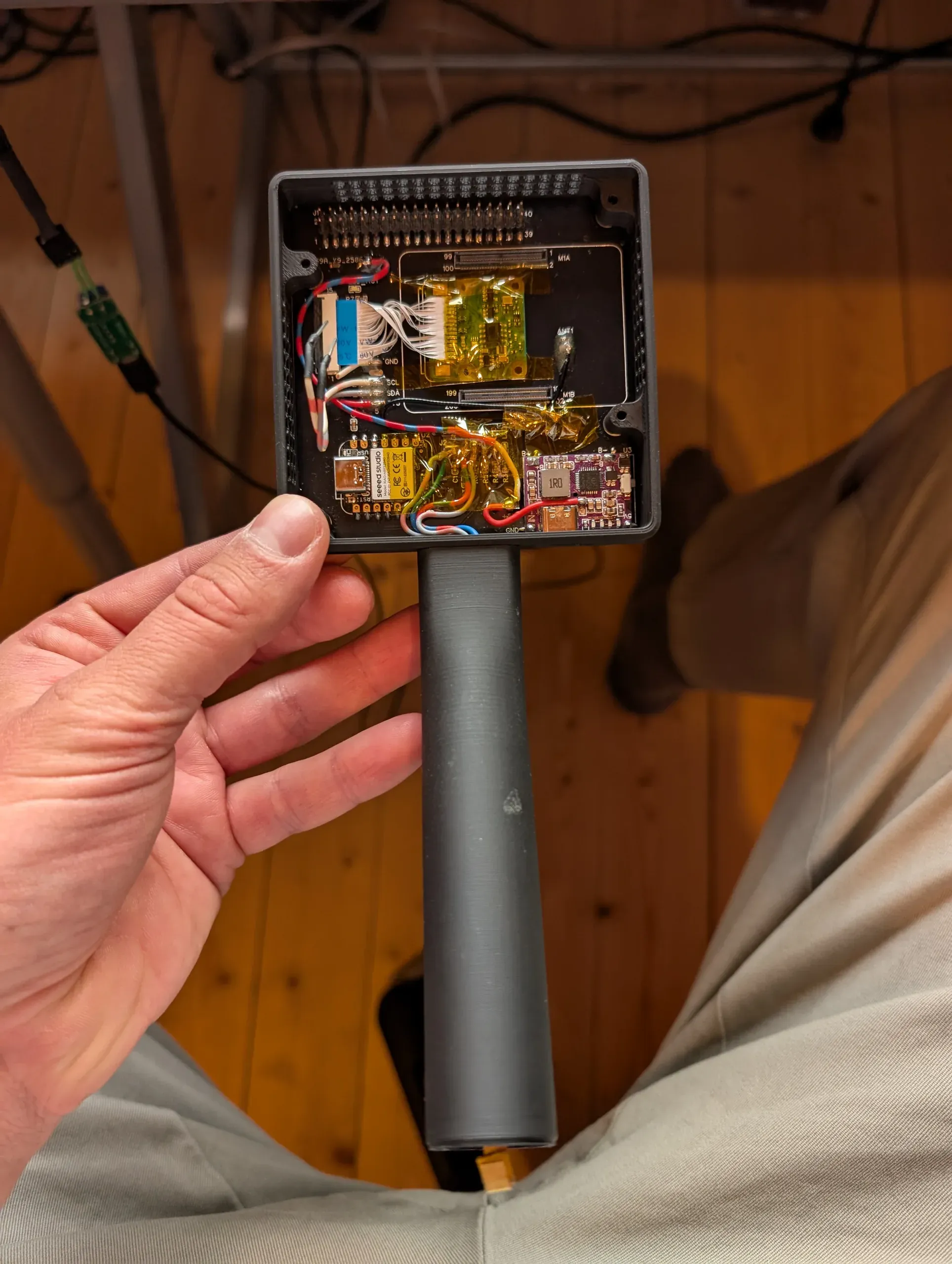 A hand holds the opened version 2: inside the square body sit the custom circuit board, a ribbon-cable adapter under amber tape, a small microcontroller and a purple charging board, with the long grip below