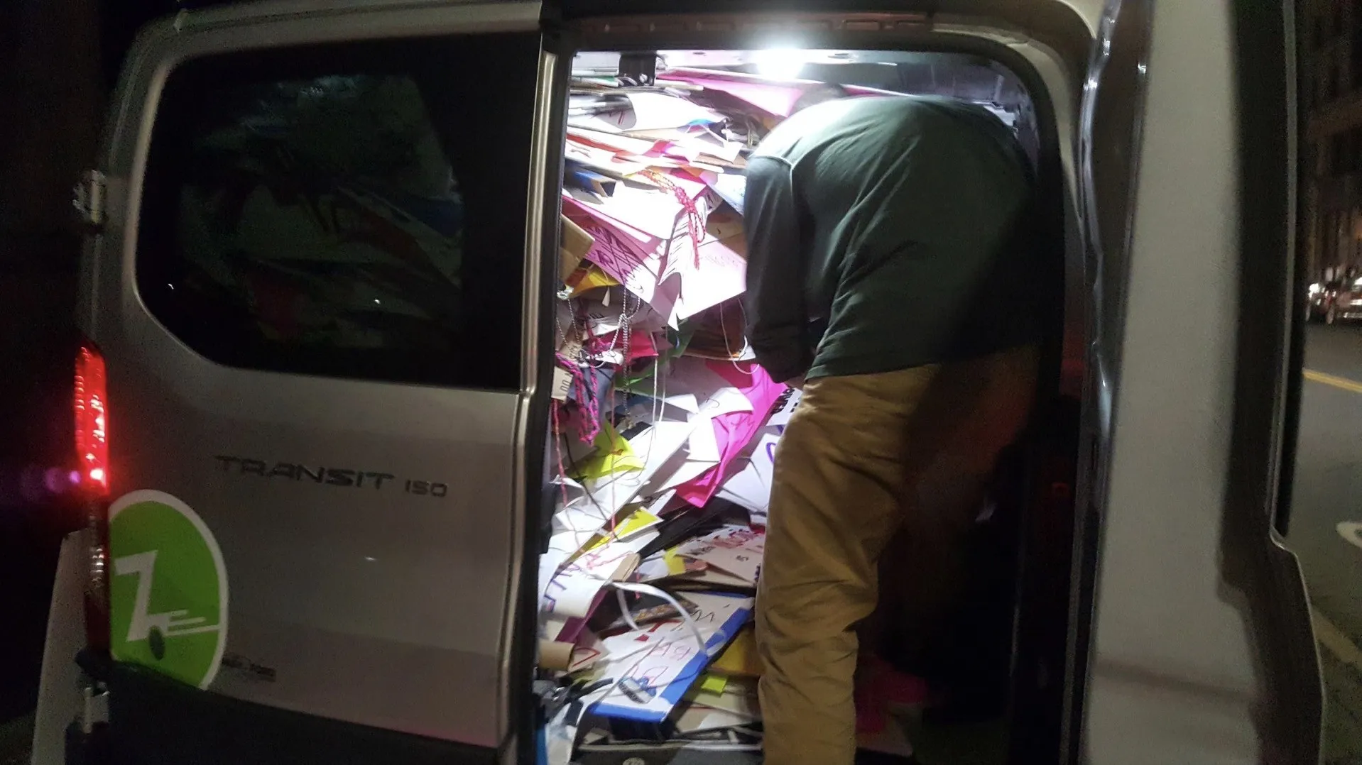 A man leans into the side door of a silver Ford Transit van at night, its cargo space packed to the roof with a jumble of pink, white and cardboard signs, some still trailing strings