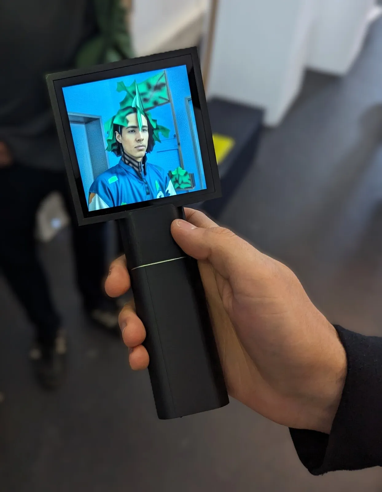 A hand holds Transferscope out; its screen shows a young man in a blue jacket whose hair has turned into green paper leaves.