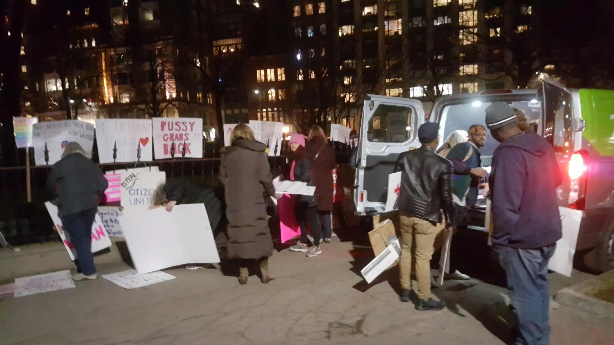 At night, volunteers in winter coats gather signs from the fence and carry them to a rented van with its back doors open, under the lit windows of high-rise buildings