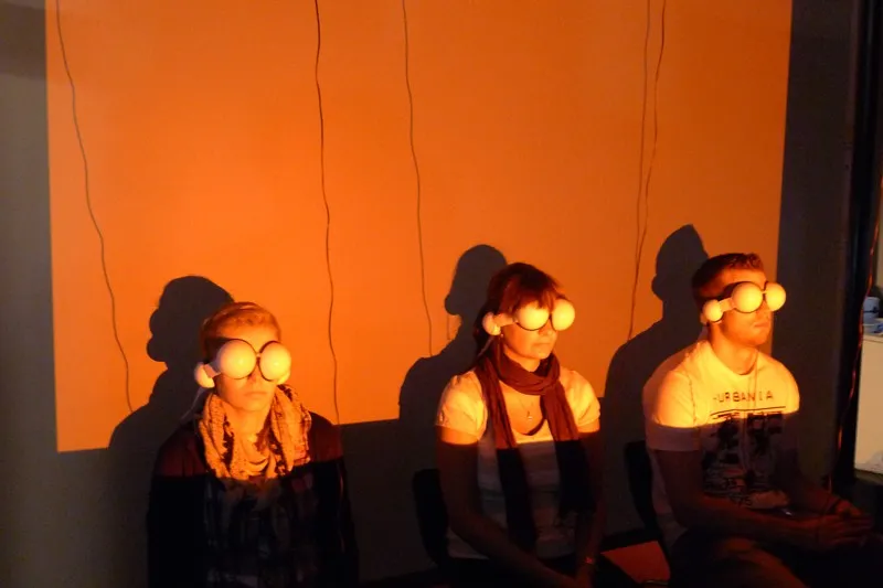 Three volunteers sit against an orange-lit wall, each wearing a diffuser mask with a white sphere over each eye and headphone shells at the ears, their faces lit by the projector